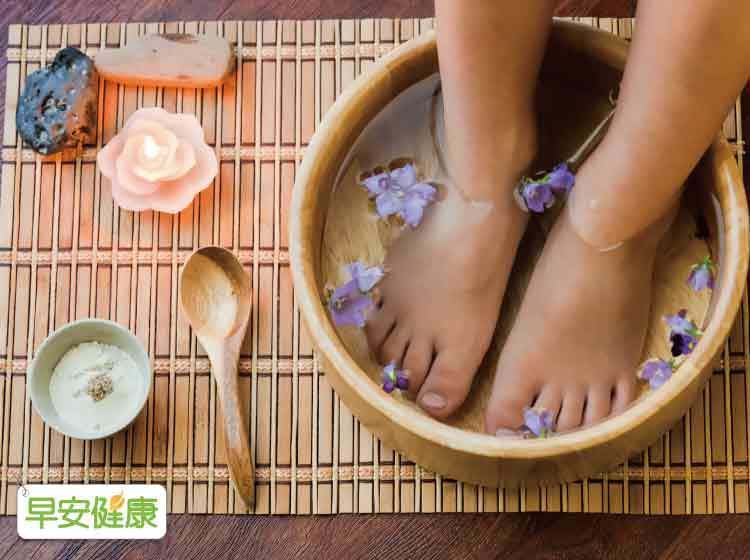 A person soaks their feet in a wooden bowl of water with flower pedals and a lit candle and herb powder to the side.